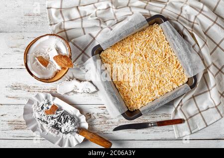 No-Bake Vanille Kokosnuss Riss quadratische Kuchen in einer Auflaufform auf einem rustikalen Holztisch mit Zutaten, vegane zuckerfreie Rezept, flach legen, horizontal vi Stockfoto