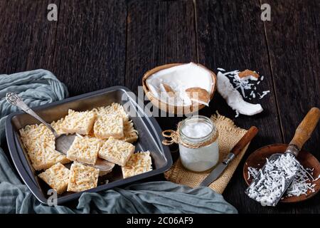 Nahaufnahme von No-Bake Kokosraspeln in einer Backform auf einem rustikalen Holztisch mit Bio-Kokosöl im Glas, horizontale Ansicht von oben Stockfoto