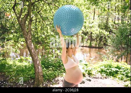 Schöne junge schwangere Frau tun Yoga-Training mit Fitness Pilates Ball im Park im Freien. Stehen und Halten mit beiden Händen. Aktive Zukunft Stockfoto