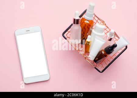 Online Kosmetik Store Konzept. Warenkorb mit Kosmetik und Handy mit leerem weißen Bildschirm Mockup auf rosa Hintergrund. Kaufen Sie ein Beauty-Produkt Stockfoto