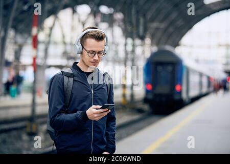 Junger Mann mit Kopfhörern, der Musik am Bahnhof mit dem Telefon gegen den Zug hört. Stockfoto