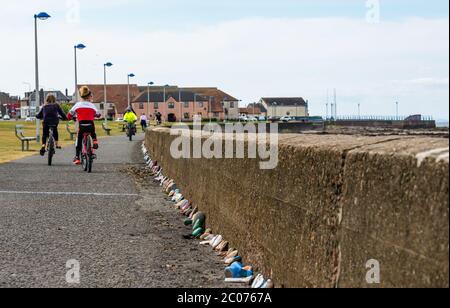 Port Seton, East Lothian, Schottland, Großbritannien. Juni 2020. Covid-19 Pandemiesymbole von Einheimischen: Eine hunderte Meter lange Linie an der Strandpromenade mit bunten und kreativen handbemalten Steinen mit inspirierenden Botschaften. Es scheint ein Phänomen zu sein, das sich in den Städten und Dörfern von East Lothian verbreitet. Menschen auf Fahrrädern radeln auf dem Fußweg an der Steinlinie vorbei, darunter zwei Mädchen auf Fahrrädern Stockfoto