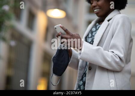 Eine Gesichtsmaske aus Stoff und ein Handy in der Hand einer glücklichen schwarzen Person. Gesichtsmasken werden verwendet, um die Ausbreitung von Coronavirus COVID-19 Pandemie zu stoppen Stockfoto