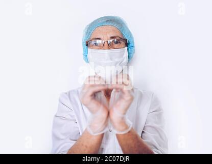 Ältere ältere Frau Arzt oder Krankenschwester zeigt Herzzeichen in einem weißen medizinischen Mantel, Handschuhe, Gesichtsmaske tragen persönliche Schutzausrüstung isoliert. Heilen Stockfoto