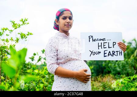 Eine junge schwangere Frau, die mit einer Plakatbotschaft an 'Bitte helfen Sie Ihrer Erde' steht. Eine schwangere Frau mit einer Plakatnachricht in der Coronavirus epid Stockfoto