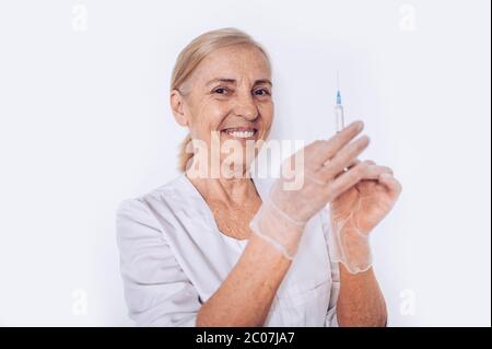 Ältere lächelnde reife Frau Arzt oder Krankenschwester mit Spritze in einem weißen medizinischen Mantel und Handschuhe tragen persönliche Schutzausrüstung isoliert. Gesundheitswesen Stockfoto