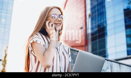 Rothaarige Unternehmer mit Brillen und Sommersprossen sitzen draußen in der Stadt und arbeiten am Laptop, während eine Telefondiskussion Stockfoto