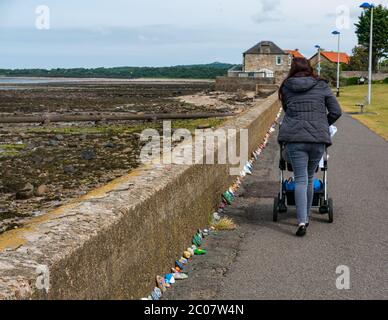 Port Seton, East Lothian, Schottland, Großbritannien. Juni 2020. Covid-19 Pandemiesymbole von Einheimischen: Eine hunderte Meter lange Linie an der Strandpromenade mit bunten und kreativen handbemalten Steinen mit inspirierenden Botschaften. Es scheint ein Phänomen zu sein, das sich in den Städten und Dörfern von East Lothian verbreitet. Eine Frau schiebt einen Buggy auf dem Fußweg an der Steinreihe vorbei Stockfoto