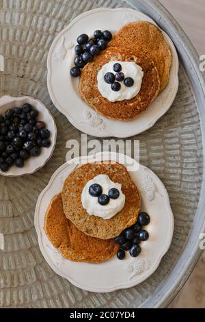 Draufsicht auf die Haferflocken Pfannkuchen serviert in zwei weißen rustikalen Teller mit Joghurt und Heidelbeeren auf einem runden Metall-Tablett Stockfoto