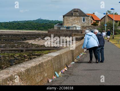 Port Seton, East Lothian, Schottland, Großbritannien. Juni 2020. Covid-19 Pandemiesymbole von Einheimischen geschaffen: Eine Linie Hunderte von Fuß lang an der Strandpromenade von bunten und kreativen handbemalten Steinen mit inspirierenden Botschaften, die die Aufmerksamkeit der Passanten anziehen. Es scheint ein Phänomen zu sein, das sich in den Städten und Dörfern von East Lothian verbreitet. Ein älteres Paar hält an, um die Steinreihe zu betrachten Stockfoto