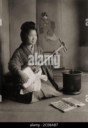 [ 1910 Japan - Shamisen spielen ] - EINE japanische Frau, die den Shamisen spielt, ein traditionelles japanisches Musikinstrument mit drei Saiten. Ihr Liederbuch liegt auf dem Boden. Silberdruck mit Vintage-Gelatine aus dem 20. Jahrhundert. Stockfoto