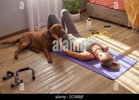 Junge Hündin, die nach dem Training zu Hause mit Hund in der Morgensonne auf der Matte liegt. Stockfoto