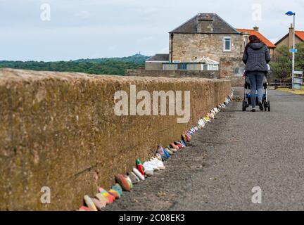 Port Seton, East Lothian, Schottland, Großbritannien. Juni 2020. Covid-19 Pandemiesymbole von Einheimischen: Eine hunderte Meter lange Linie an der Strandpromenade mit bunten und kreativen handbemalten Steinen mit inspirierenden Botschaften. Es scheint ein Phänomen zu sein, das sich in den Städten und Dörfern von East Lothian verbreitet. Eine Frau schiebt einen Buggy auf dem Fußweg an der Steinreihe vorbei Stockfoto
