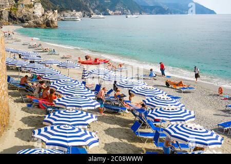 Der Sandstrand von Monterosso, Cinque Terre Italien, mit Sonnenschirmen, Sonnenanbeter, das Ligurische Meer und Boote entlang der felsigen Küste. Stockfoto