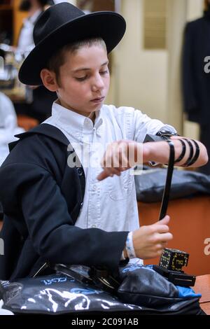 Ein orthodoxer jüdischer Junge legt seinen Tefillin vor dem morgendlichen Gebetsgottesdienst in einer Synagoge in Crown Heights, Brooklyn, New yourk an. Stockfoto