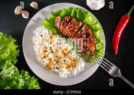 Teriyaki Huhn mit Sesam, Salat und Reis auf einem weißen Teller. Gericht mit einer Gabel auf schwarzem Hintergrund Stockfoto