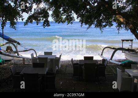 Leere Terrasse ohne Touristen und Jukung Boote an sonnigen Tagen in Bali, Indonesien. Tische und Stühle stehen vor Booten einer Strandbar Stockfoto