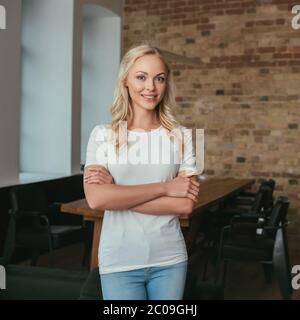 Glückliche blonde Frau lächelt vor der Kamera, während sie zu Hause mit verschränkten Armen steht Stockfoto