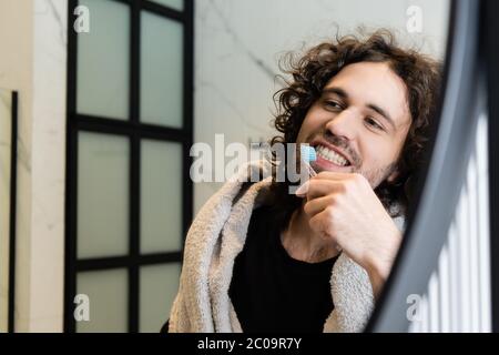 Selektiver Fokus von lockigen Mann mit Handtuch auf den Schultern putzen Zähne im Bad Stockfoto