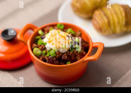 Frische hausgemachte Schüssel mit Chili con Carne mit Bohnen, saurer Sahne, geriebenem Cheddar-Käse und grünen Zwiebeln. Serviert mit hasselback Kartoffeln auf der Seite. Stockfoto