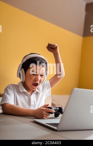 KIEW, UKRAINE - 22. APRIL 2020: Emotionaler asiatischer Junge, der Videospiel mit Joystick und Laptop auf Quarantäne spielt Stockfoto