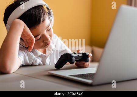 KIEW, UKRAINE - 22. APRIL 2020: Schläfriger asiatischer Junge, der Videospiel mit Joystick und Laptop auf Quarantäne spielt Stockfoto