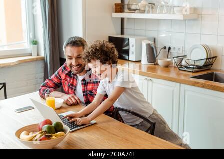Fröhlicher Junge mit Laptop in der Nähe Freelancer Vater und Frühstück auf dem Tisch Stockfoto