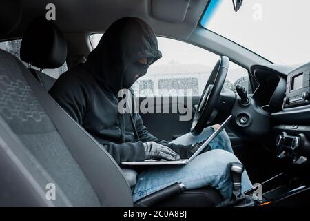 Seitenansicht des Räubers in Sturmhaube und Lederhandschuhe mit Laptop während im Auto sitzen Stockfoto