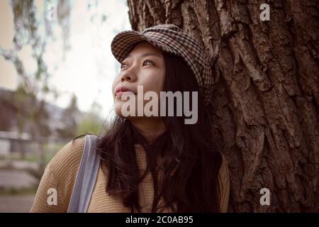 Attraktive asiatische weibliche College Student Blick in die Entfernung während Schiefen gegen EINEN Baum Stockfoto