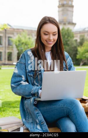 Fröhliche Student auf Bank sitzen und mit Laptop in der Nähe von Büchern, Online-Studie Konzept Stockfoto