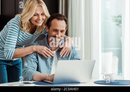 Selektiver Fokus der lächelnden Frau umarmen Ehemann mit Laptop in der Küche Stockfoto