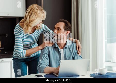 Selektiver Fokus der lächelnden Frau umarmen schönen Mann mit Laptop auf Küchentisch Stockfoto
