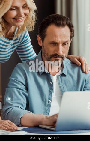 Selektiver Fokus der lächelnden reifen Frau umarmen Ehemann mit Laptop am Tisch Stockfoto