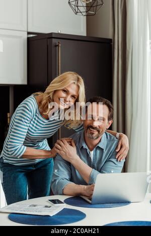 Selektiver Fokus der reifen Frau lächelt auf Kamera, während umarmen Mann in der Nähe von digitalen Geräten auf Küchentisch Stockfoto