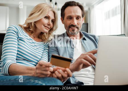 Selektiver Fokus des lächelnden Mannes zeigt auf Laptop in der Nähe der Frau mit Kreditkarte im Wohnzimmer Stockfoto