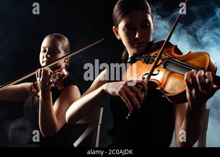 Schöne Musiker spielen auf Geigen auf dunkler Bühne mit Rauch Stockfoto