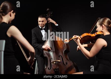 Trio von glücklichen Musikern, die auf Kontrabass spielen und Violinen isoliert auf Schwarz Stockfoto