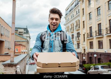 Selektiver Fokus der fröhlichen Kurier hält Pizzakisten mit städtischen Straße im Hintergrund Stockfoto