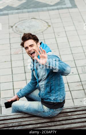 Hohe Winkelansicht des positiven Mannes in Ohrhörern, die Smartphone halten und Hand auf Bank winken Stockfoto