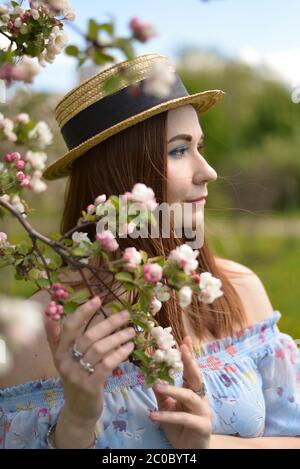 Ein schönes Mädchen in einem blauen Kleid und einem Stroh hut hält einen blühenden Zweig eines Apfelbaums und Schaut in die Ferne Stockfoto