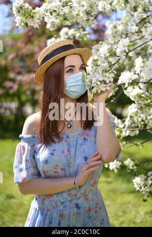 Ein schönes Mädchen in einer medizinischen Maske, blauem Kleid und Strohhut steht bei einem blühenden weißen Apfelbaum im Garten Stockfoto