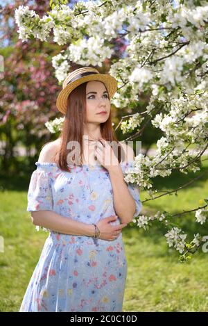 Ein schönes Mädchen in einem blauen Kleid und einem Stroh hut steht bei einem blühenden weißen Apfelbaum im Garten Stockfoto