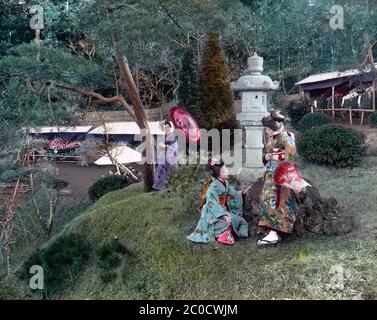 [ 1900er Japan - Japanischer Garten ] - zwei junge Frauen sitzen vor einer Steinlaterne in einem Garten in Yokohama, Präfektur Kanagawa. Eine dritte Frau, die einen Sonnenschirm hält, steht im Rücken. Der private Garten befand sich auf dem Gelände einer Villa in Nogeyama, die einer reichen Handelsfamilie gehörte. Zweimal im Jahr wurde der Garten für die Öffentlichkeit geöffnet und die Menschen würden Schlange stehen, um die Pflaumenblüte im Frühjahr und die Chrysanthemen, sichtbar im Hintergrund dieser Szene, im Herbst zu bewundern. Vintage-Glasrutsche aus dem 20. Jahrhundert. Stockfoto