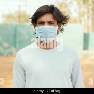 Junger Mann mit medizinischer Maske Blick in die Kamera, während Wind weht seine Haare während heißen sonnigen Tag - Konzept der Maske im Freien zu stoppen tragen Stockfoto