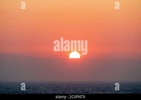 Sonnenaufgang über dem Meer vor der Broadstairs Küste in Kent, England. Teleaufnahme der Sonne, die über einer Schicht von trüben Wolken am Horizont über dem Meer aufgeht. Roter Himmel dreht sich oben zu gelb. Die Sonne teilweise durch die Wolke versteckt. Stockfoto