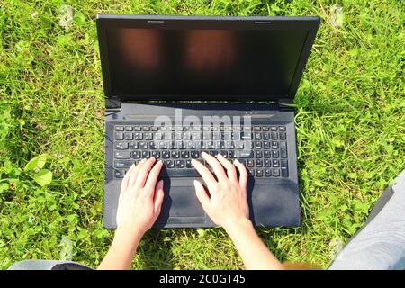 Das Mädchen sitzt auf dem grünen Gras auf dem Rasen und hält einen schwarzen Laptop auf dem Schoß. Nahaufnahme des Laptops. Stockfoto