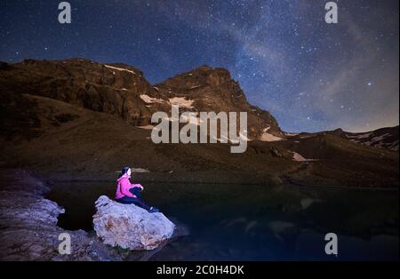 Seitenansicht einer jungen Frau, die auf einem großen Stein am Seeufer sitzt. Herrliche Aussicht auf majestätische felsige Hügel Matterhorn mit Dame Reisenden unter Nacht Sternenhimmel. Konzept von Reisen, Wandern und Nacht. Stockfoto