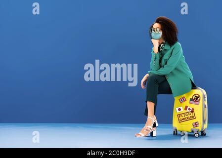 Nachdenkliche afroamerikanische Frau sitzt in medizinischer Maske auf Reisetasche auf blauem Hintergrund Stockfoto
