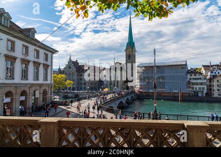 Zürich, Schweiz - 6. Oktober 2018: Blick auf die Kirche Fraumünster. Besucher besuchen Sehenswürdigkeiten und wandern über die Münsterbrücke über einen Fluss. Stockfoto