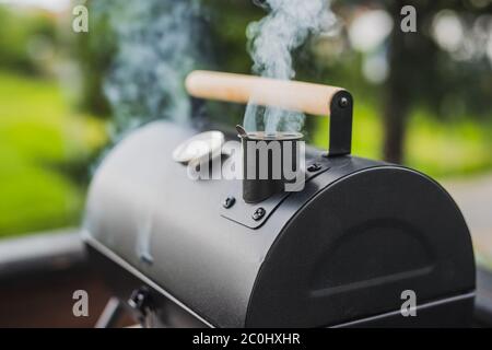 Rauch kommt aus einem Smokestack eines kleinen schwarzen Smoker Grill oder Grill auf grünem Hintergrund. Moderner Rauchergrill auf der Veranda, keine Leute gesehen. Stockfoto